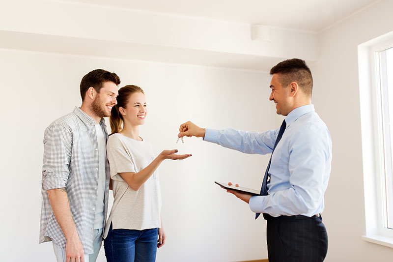Home 210 A man in a suit hands keys to a smiling couple inside a bright, empty room, possibly signifying the purchase or rental of a new home. The woman reaches out to accept the keys.