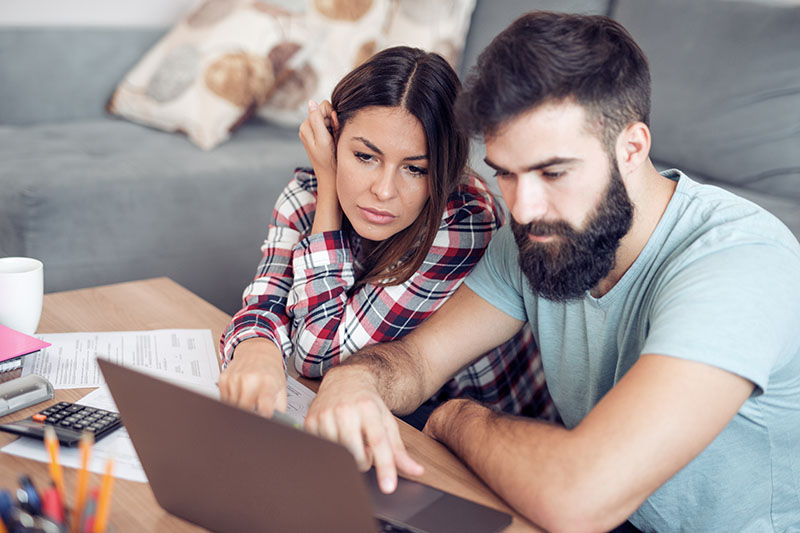 Home 213 A couple sits together at a table, looking concerned while reviewing documents and using a laptop. Papers, a calculator, and a mug are on the table, suggesting they are managing finances or budgeting at home.