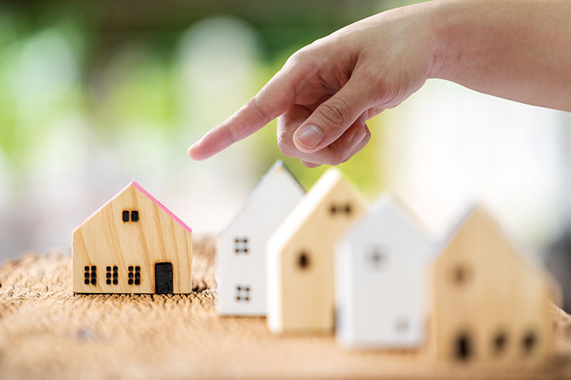 Home 215 A hand points toward a small wooden house model with a pink roof among other similar house models placed on a textured surface, suggesting the concept of choosing a home or property selection.