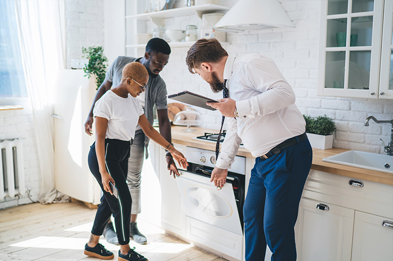 Home 232 A man in a suit shows a couple an oven in a modern white kitchen. The couple looks closely at the appliance while the man holds a clipboard and gestures toward the open oven door.