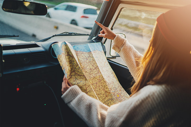 Home 220 A person in a sweater and hat sits in a car passenger seat, holding a map while pointing out the windshield, suggesting they are navigating or giving directions during a road trip. Sunlight streams through the window.