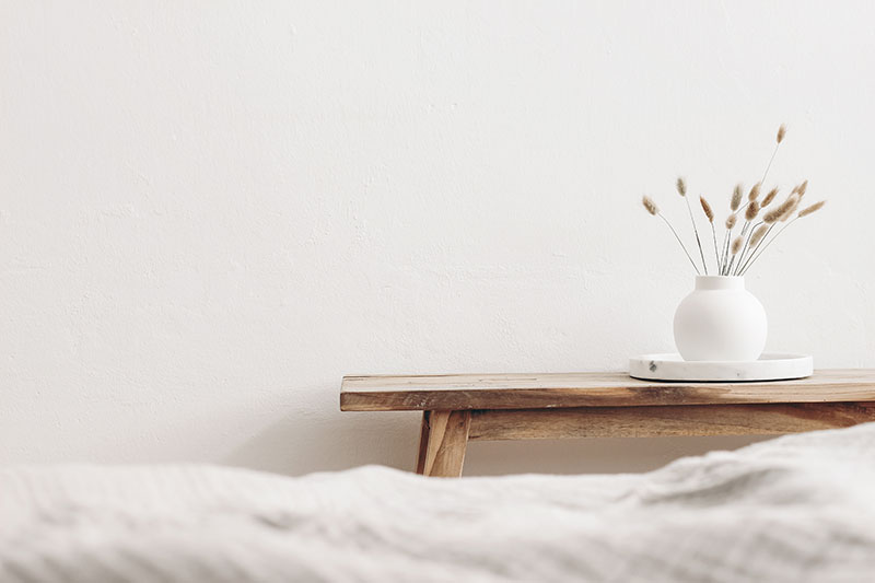 Home 238 A minimalist scene with a wooden bench against a white wall, holding a round white vase filled with dried grass on a white tray. The foreground shows the edge of a soft, light-colored blanket.