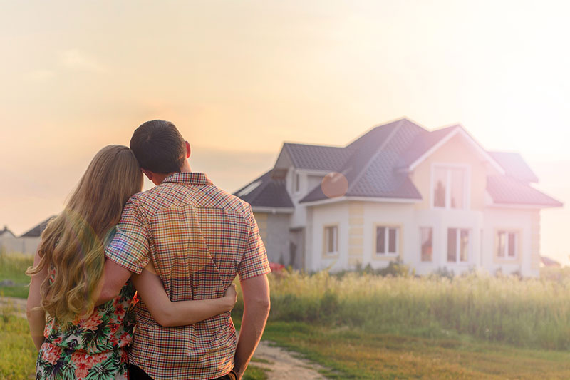 Home 227 A couple stands outdoors with their arms around each other, looking at a modern house in the distance under a bright, hazy sky.