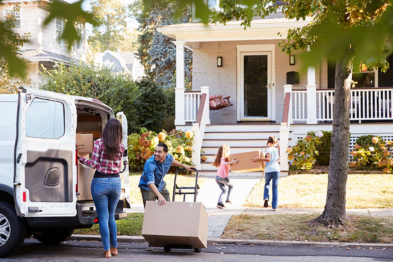 Home 241 A family is moving into a new house; a man wheels a box on a dolly, a woman loads a van, and two children carry boxes up the walkway. The house has a porch and is surrounded by trees and bushes.