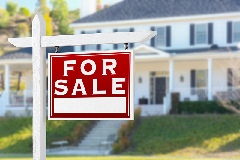 Home 236 A red and white For Sale sign is posted in front of a blurred, large two-story suburban house with white siding, black shutters, and a front porch.