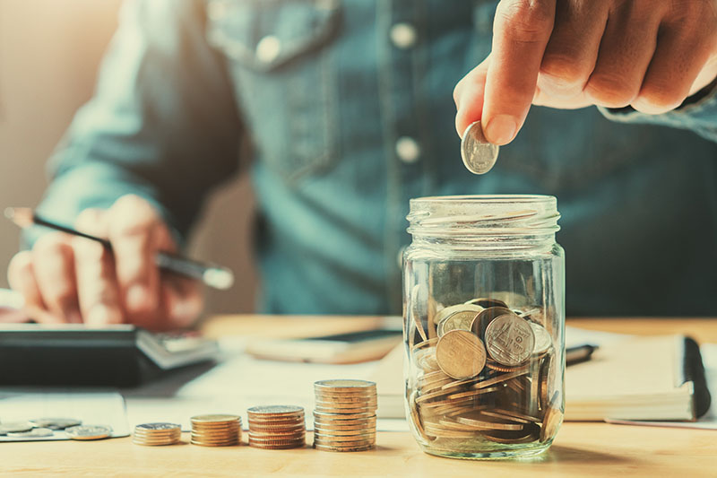 Home 224 A person in a denim shirt drops a coin into a glass jar filled with coins. Stacks of coins, a notebook, and a hand using a calculator are visible on the table in the background.
