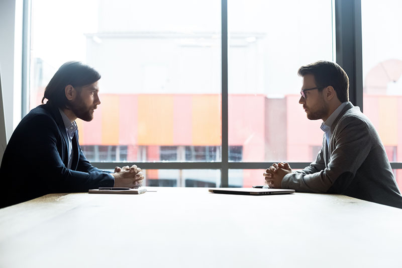 Home 239 Two men sit across from each other at a table in an office setting, facing each other with serious expressions, hands clasped, and large windows in the background letting in natural light.