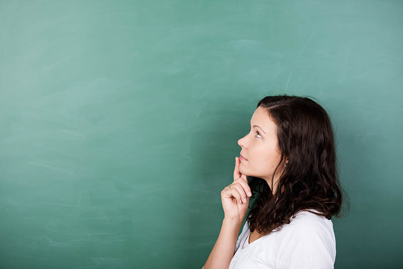 Home 225 A woman with long brown hair, wearing a white shirt, stands in profile against a green chalkboard, looking thoughtful with her finger resting on her chin.