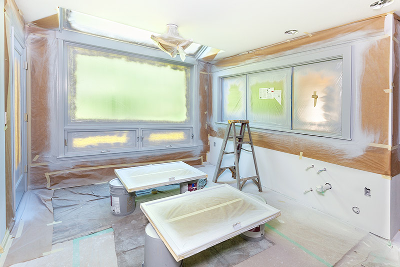 Rehab Loan 1 A room prepared for painting with plastic coverings on the floor and windows, two cabinet doors on sawhorses, a step ladder, and paint supplies scattered around. The walls and woodwork are primed and ready for paint.