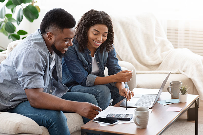 Home 202 A smiling couple sits on a couch, reviewing financial documents and using a laptop and calculator on a coffee table, appearing focused and happy while managing their finances at home.