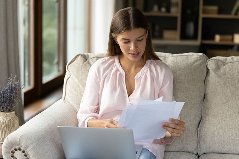 Home 204 A woman sits on a couch with a laptop, looking at papers in her hands. She wears a light pink blouse and appears to be reviewing documents in a bright, cozy living room.