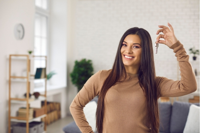 Home 200 A smiling woman with long brown hair stands in a bright living room, holding up a key in her right hand, suggesting she has just moved in or bought a new home.