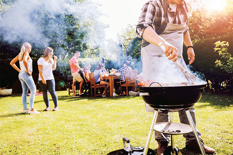 Home 205 A person grilling food on a barbecue in a sunny backyard while a group of people socialize and sit at a table in the background. Smoke rises from the grill, and trees and grass surround the scene.