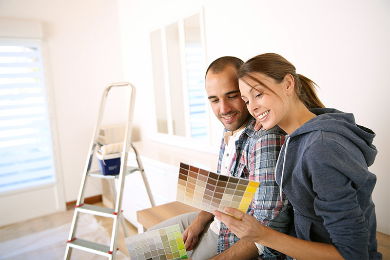 Home 207 A smiling couple sits in a bright room, looking at paint color swatches. A ladder and painting supplies are in the background, suggesting they are planning to redecorate or paint the room together.