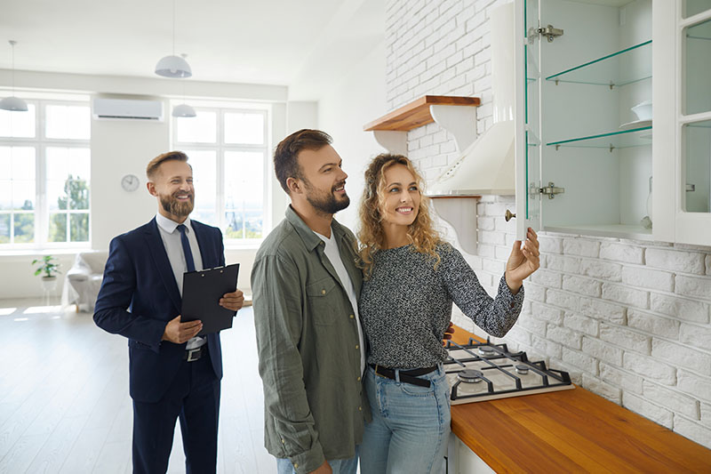 Home 206 A smiling couple examines a glass kitchen cabinet in a modern, bright kitchen while a real estate agent in a suit stands nearby holding a clipboard.