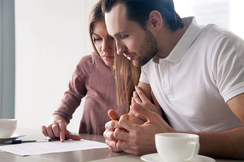 Home 208 A couple sits closely together at a table, reading and discussing a document. One person points at the paper with a pen, while both look focused and serious. Two cups are on the table in front of them.