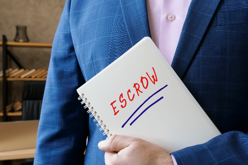 Home 195 A person in a blue suit holds a notebook with the word ESCROW written in red marker, underlined in blue. The background is an office setting with shelves and hangers.