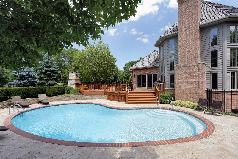 Home 197 A kidney-shaped swimming pool with a brick border in a backyard, surrounded by a patio, wooden deck, lounge chairs, and a large brick house with tall windows and a chimney. Lush greenery and trees are in the background.