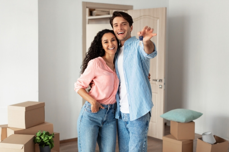 Home 198 A smiling couple stands close together in a room with moving boxes, with the man pointing playfully towards the camera. The room is bright and has an open door and a plant in the background.