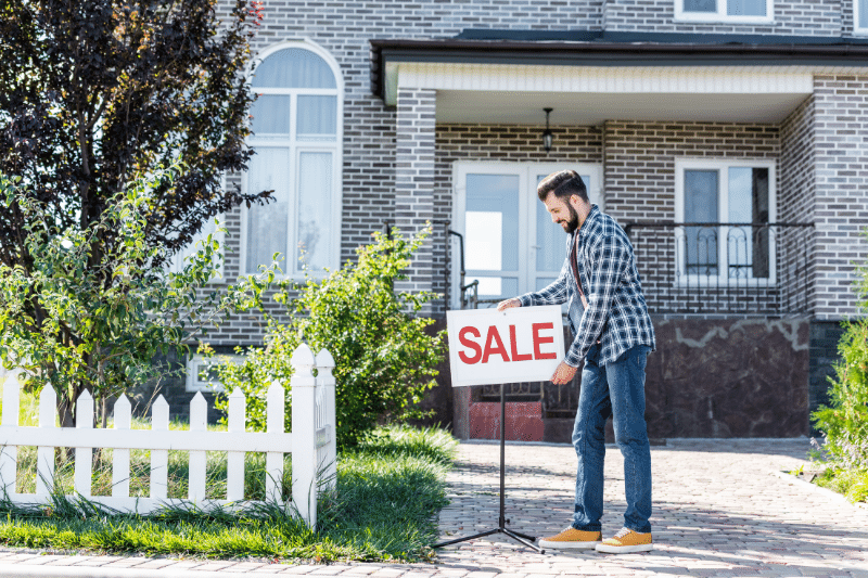 Home 196 A man places a SALE sign in front of a modern brick house with a white fence and green lawn.