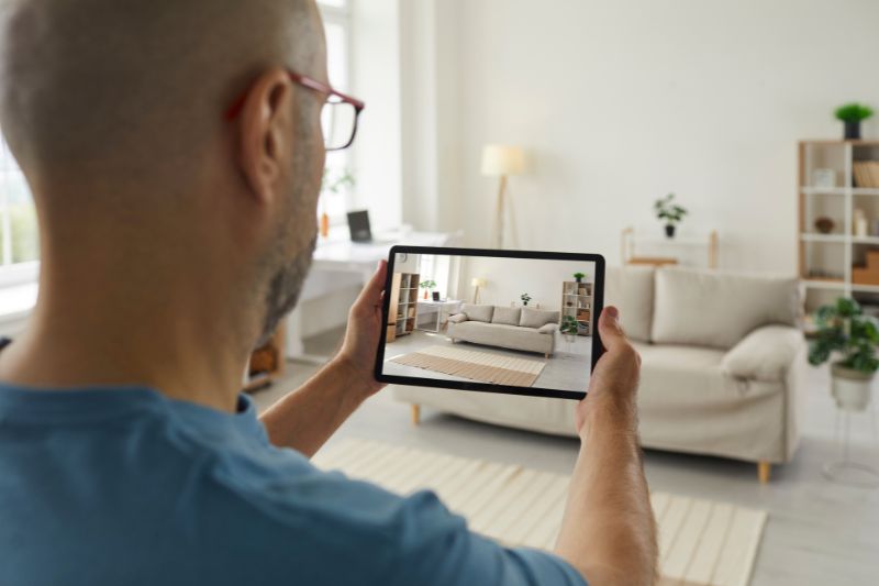 Home 190 A person holding a tablet takes a photo of a modern living room with a beige sofa, a rug, and plants. The room appears bright and neatly decorated with natural light from the windows.