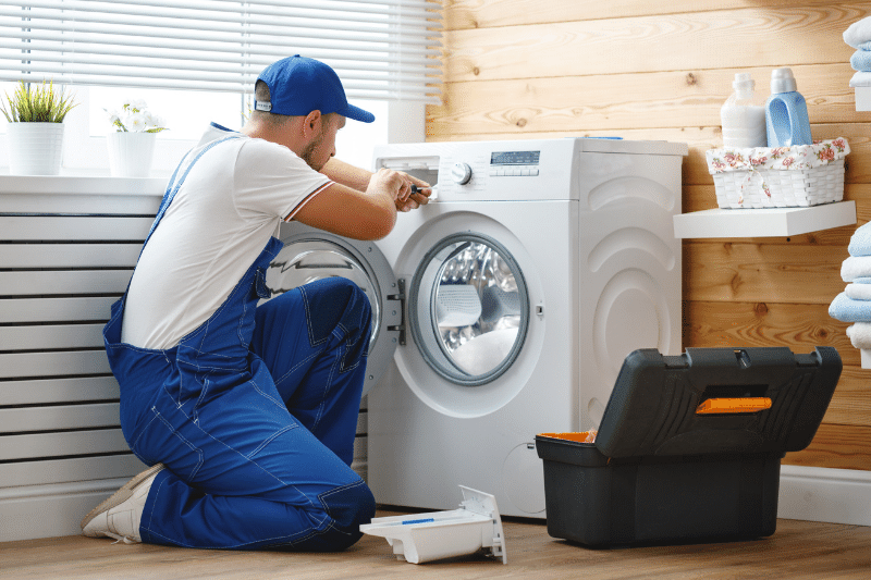 Home 193 A repairman in blue overalls kneels in front of a washing machine, using a tool to fix it. An open toolbox sits nearby in a bright laundry room with folded towels and cleaning supplies.