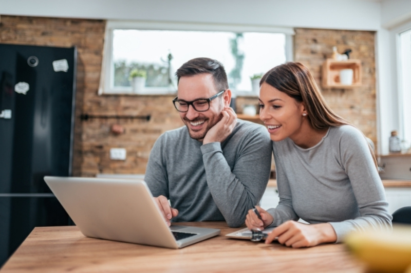 Home 186 A smiling couple sits at a kitchen table looking at a laptop. The man is wearing glasses and both are dressed casually. The woman holds a credit card and a notepad. The kitchen has a brick wall and a window in the background.