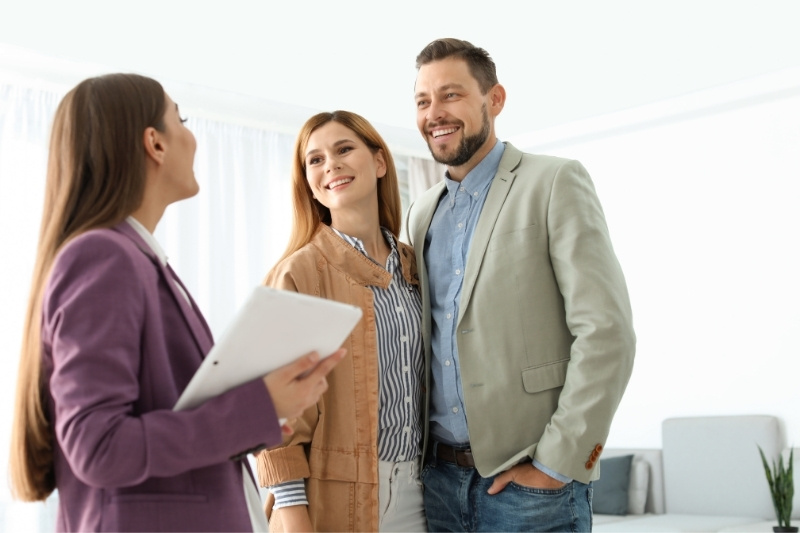 Home 187 A woman holding a tablet smiles while talking to a happy couple dressed in business casual attire, standing together in a bright, modern room.
