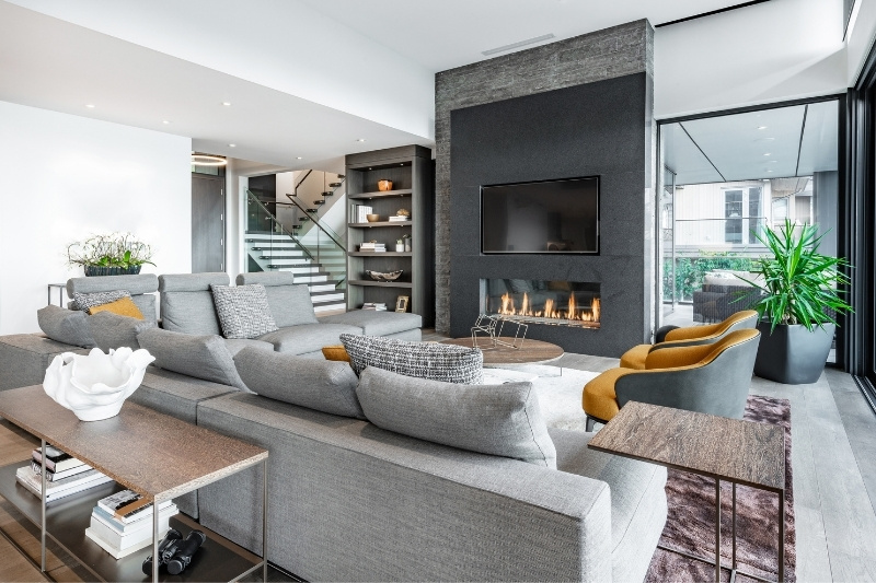 Home 184 Modern living room with a large gray sectional sofa, built-in shelves, a fireplace under a wall-mounted TV, two mustard chairs, a large window, and green indoor plants, decorated in neutral tones.