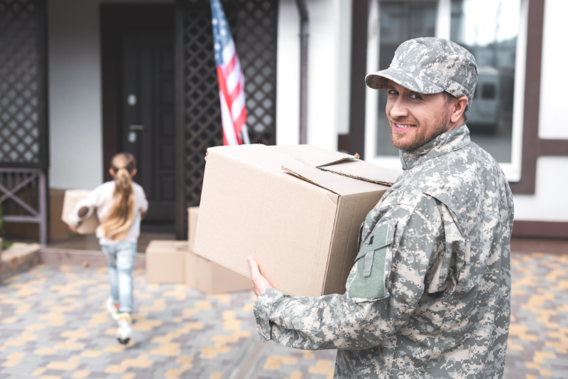 Home 181 A man in military uniform smiles while holding a cardboard box outside a house. An American flag is near the door, and a young girl carrying a box walks ahead of him toward the house.