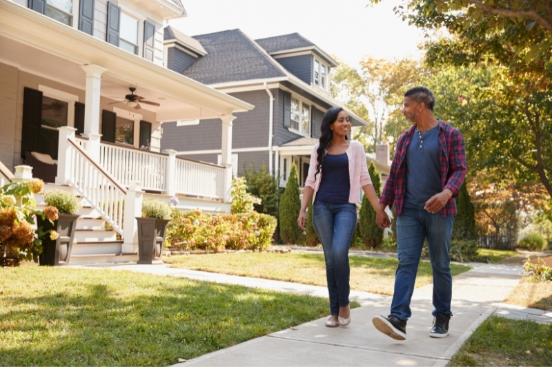 Home 183 A couple walks hand in hand on a sidewalk in a suburban neighborhood, passing by houses with front porches and gardens on a sunny day.