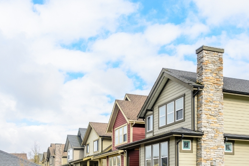 Home 185 A row of modern suburban houses with varied siding colors and stone chimneys, seen under a partly cloudy blue sky.