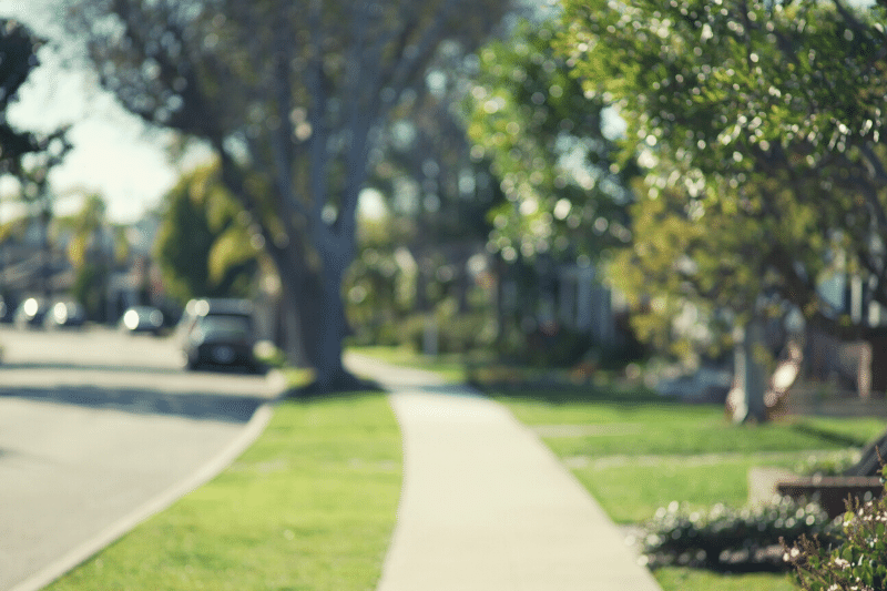 Home 182 A sunlit suburban sidewalk lined with green grass and trees, with houses and a parked car visible in the background. The image is softly out of focus, creating a peaceful, dreamy atmosphere.