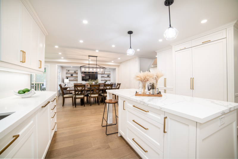 Home 177 Spacious modern kitchen with white cabinetry, gold hardware, marble countertops, wooden floors, and pendant lights. A kitchen island with stools and decorative accents faces a dining area and living space in the background.