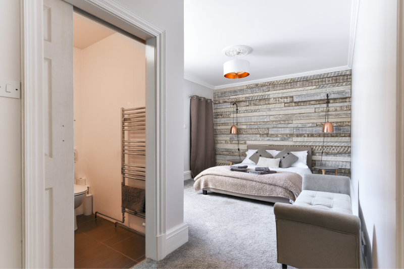 Home 178 A modern bedroom with a rustic wood accent wall, double bed, neutral bedding, wall lamps, gray bench, and an open door leading to a small en-suite bathroom with a towel warmer.