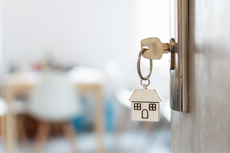 Home 175 A key with a house-shaped keychain is inserted into a door lock, with a blurred interior room visible in the background.