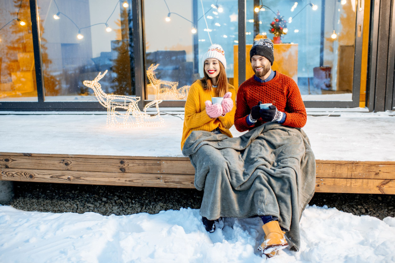 Home 176 A smiling couple in winter clothes sits on a snowy porch, wrapped in blankets and holding mugs. Festive decorations and string lights are behind them, along with a lit reindeer figure.