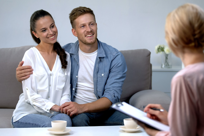 Home 138 A smiling couple sits close together on a couch, holding hands and talking to a person with a clipboard, possibly a therapist or counselor, in a comfortable room with coffee cups on a table.