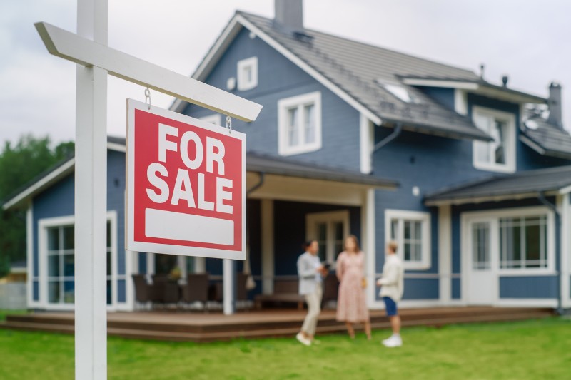 Home 174 A red and white For Sale sign is in the foreground, with a modern blue house and three people talking on the front lawn in the background.