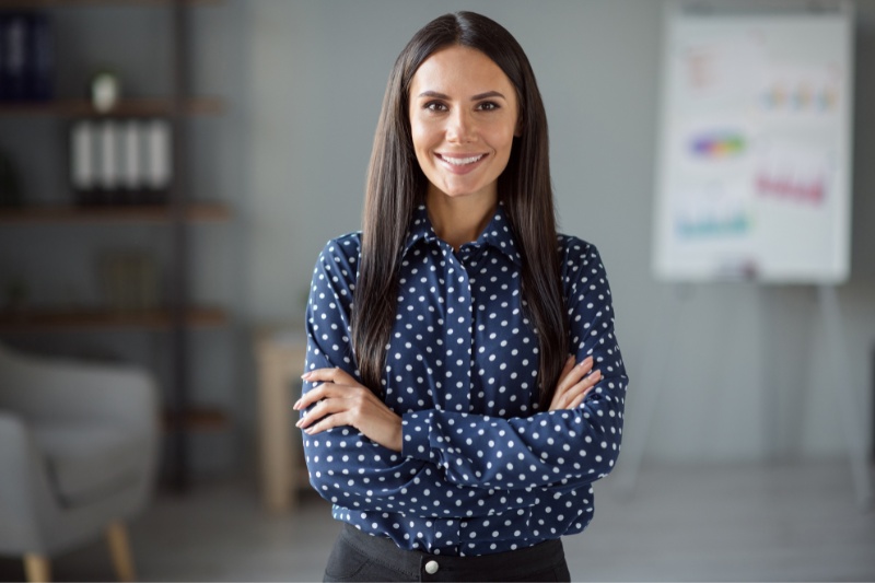 Home 132 A woman with long dark hair, wearing a navy blue polka dot shirt and black pants, stands confidently with arms crossed and smiles in an office setting. A blurred flipchart and shelves are visible in the background.