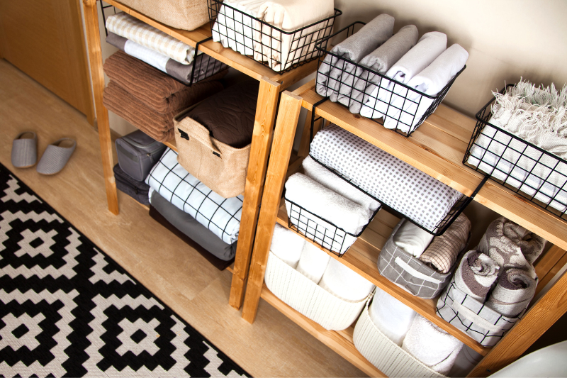 Home 135 Wooden shelves neatly organize towels, linens, and baskets in a bright room. A black-and-white geometric rug and gray slippers are on the floor, creating a tidy, cozy atmosphere.