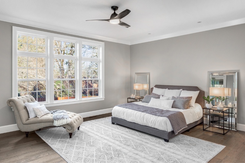 Home 133 A modern bedroom with large windows, a gray upholstered bed with white and gray bedding, matching nightstands with lamps, a tufted chaise lounge, a patterned rug, and light gray walls with wood flooring.