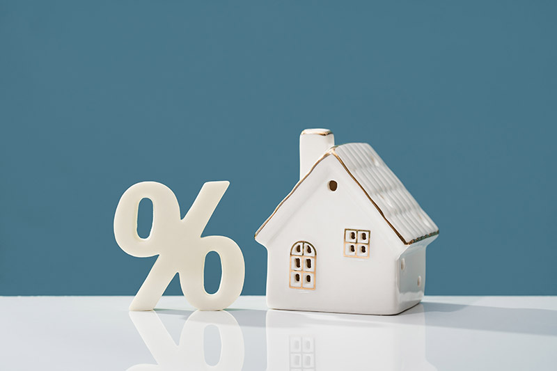 Home 134 A small ceramic house and a large white percentage symbol are placed side by side on a reflective surface, against a plain blue background.