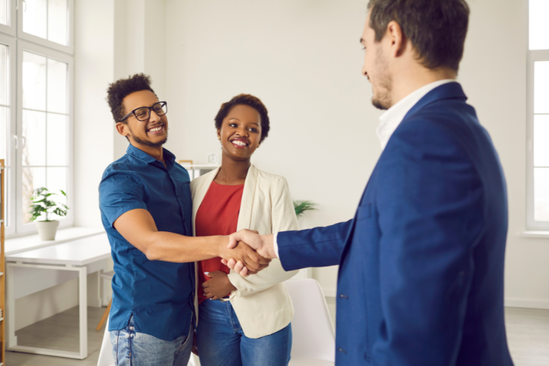 Home 128 A smiling man in a blue shirt shakes hands with a man in a suit while a woman in a white blazer stands beside them, all in a bright office setting.