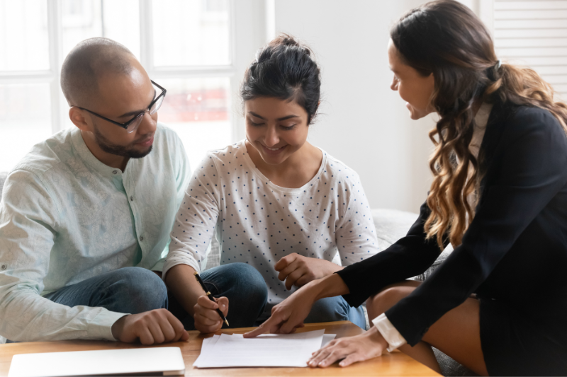 Home 129 A woman in business attire points to a document on a table while a seated man and woman listen and review the paper, appearing to discuss or sign paperwork together in a bright room.