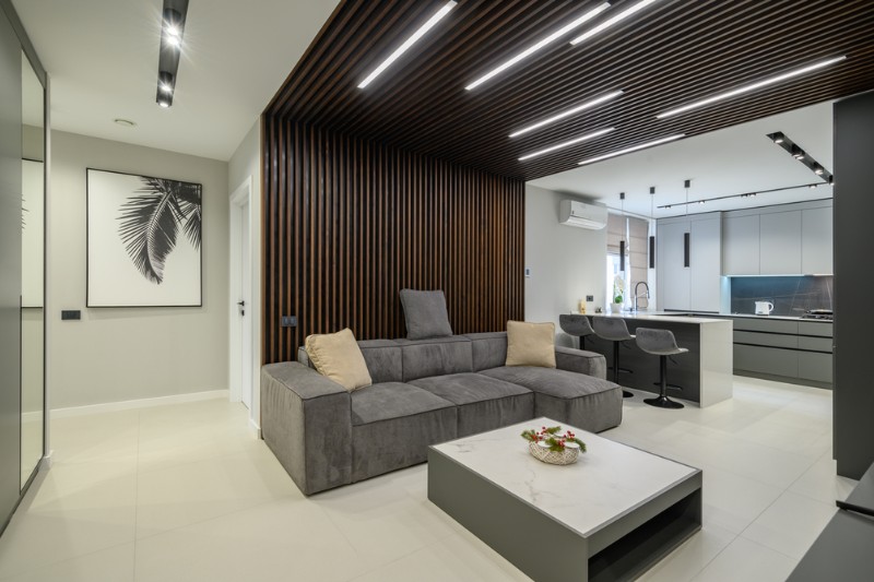 Home 124 Modern living room with a gray sectional sofa, a low white coffee table, and a wall with vertical wooden slats. The open kitchen has bar stools and sleek cabinetry. Minimalist decor with neutral tones.