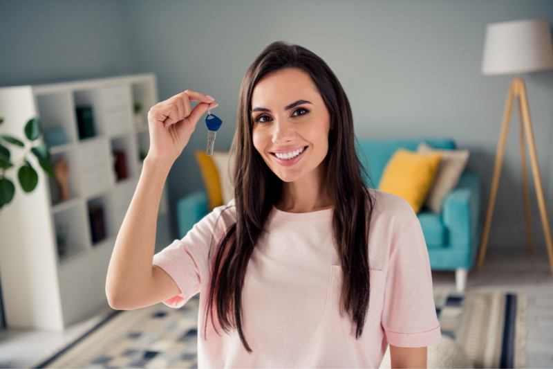 Home 123 A smiling woman with long dark hair holds up a key indoors, standing in a stylish living room with a blue sofa, yellow pillows, a bookshelf, and a floor lamp in the background.