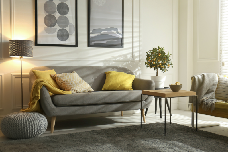 Home 126 A modern living room with a gray sofa, yellow and gray pillows, a knitted pouf, a wooden coffee table, a potted plant, floor lamp, and abstract wall art. Natural light streams through window blinds.