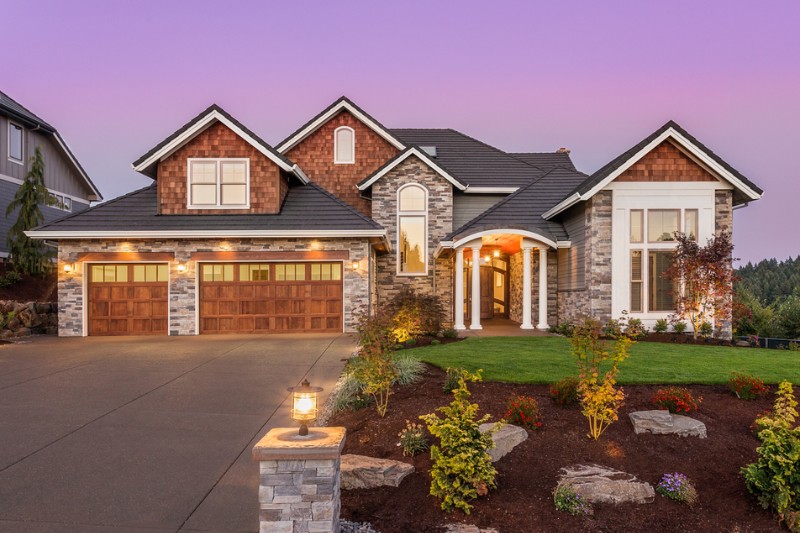 Home 125 A large, modern suburban house with stone and wood exterior, three-car garage with wooden doors, a well-manicured lawn, landscaped garden, and a driveway at sunset.