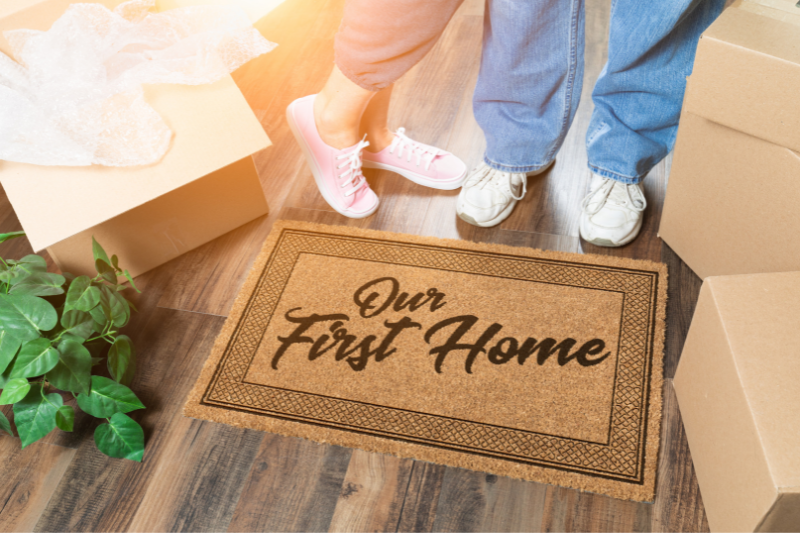 Home 122 Two people stand next to a doormat that reads Our First Home, surrounded by moving boxes and a plant on a wooden floor, with sunlight shining in.